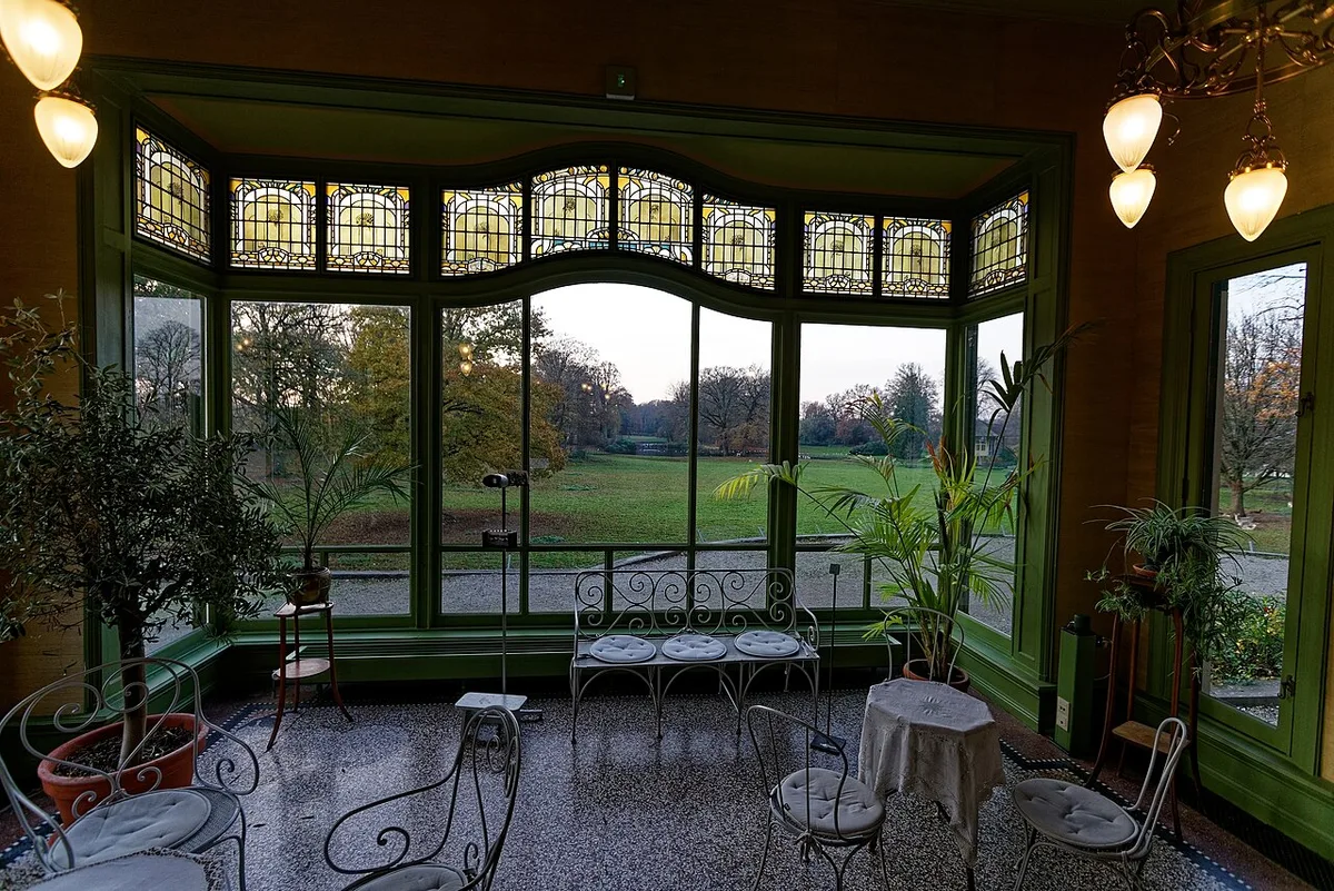 Véranda ancienne à verrière et vitraux verts, meublée de tables et de chaises en fer forgé, ouvrant sur un parc.