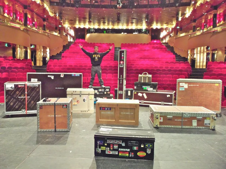 Un homme se tient parmi des caisses de matériel sur une scène, devant une salle de spectacle vide aux fauteuils rouges.